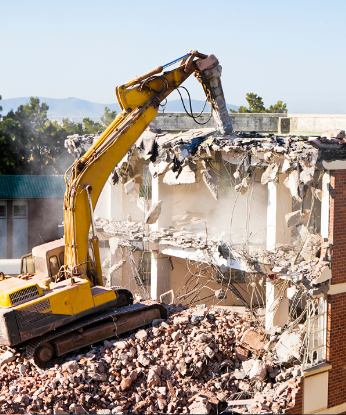 A yellow excavator digging through rubble in front of a building